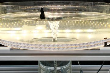 A cylindrical, transparent container stands on an open, silver-coloured metal frame. It is filled with clear water, at the centre of which a slender, funnel-shaped vortex rotates from surface to bottom. The water surface is indented at the centre and slightly rippled at the edges. Tiny air bubbles are drawn downward by the vortex, forming fine lines along the inner wall. LED light strips encircle the container, making the vortex stand out starkly against the dark surroundings. The scene has the appearance of a scientific demonstration visualising fluid dynamics.