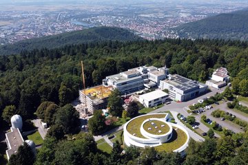 Luftaufnahme des Campus des Max-Planck-Instituts für Astronomie (MPIA) auf dem Königstuhl in Heidelberg. Im Vordergrund rechts unten ist das Haus der Astronomie zu sehen, ein modernes, weißes Gebäude in Spiralform mit begrüntem Dach. Links davon befinden sich mehrere weitere Institutsgebäude mit Flachdächern, teilweise mit Solarpaneelen, sowie ein Neubau mit Baukran und gelber Gerüstverkleidung. Im unteren linken Bereich sind zwei weiße Kuppeln kleiner Observatorien zu erkennen. Vor den Gebäuden liegen Parkplätze mit geparkten Autos. Umgeben ist der Campus von dichtem grünen Wald. Im Hintergrund erstreckt sich das Neckartal mit der Stadt Heidelberg, Flussläufen, Brücken und dem weiten, flachen Oberrheingebiet bis zum Horizont.