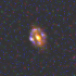Abbildung 1: Aufnahme des Weltraumteleskops Hubble von J1000+0221, der entferntesten bekannten Gravitationslinse. Das Licht des massereichen Objekt, das als Linse wirkt, benötigt 9,4 Milliarden Jahre (z = 1,53) um uns zu erreichen. Die Vordergrundgalaxie (Linsenmasse) erscheint orange gefärbt, die Hintergrundgalaxie, die in Form eines Einsteinrings vergrößert wird, bläulich.Der bläuliche Einsteinring misst nur 0,7 Bogensekunden im Durchmesser (entsprechend eines Durchmessers von 19,000 Lichtjahren am Ort der Linse). Das ist weniger als 1/2500 des Durchmessers des Vollmonds und an der Grenze desjenigen, was herkömmliche bodengebundene Teleskope überhaupt abbilden können. Das Objekt ist so klein, dass in dem Bild bereits die Pixelstruktur des Detektorchips sichtbar wird.Das Farbbild wurde aus drei separaten Aufnahmen mit zwei Teleskope an Bord des Weltraumteleskops Hubble erstellt: zwei nahinfraroten Bildern der Wide Field Camera 3 (Quelle: NASA, ESA und CANDELS) und einem Bild der Advanced Camera for Surveys (Quelle: NASA, ESA, CANDELS und COSMOS).