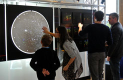 Foto einer Kunstausstellung von SDSS Artist-in-Residence Tim Fitzpatrick in St Andrews, Schottland, am 7. November 2015. Freiwillige und Besucher untersuchen eine hinterleuchtete SDSS-Platte auf der linken Seite und Kunst, die von astronomischen Spektren inspiriert ist, auf der rechten Seite.