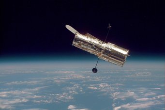 The Hubble Space Telescope drifts 353 miles (569 km) above the Earth's surface, where it can avoid the atmosphere and clearly see objects in space. The space telescope is floating in space. In the background, the upper half of the image shows black space. The lower half shows the bluish shimmering earth with clouds.