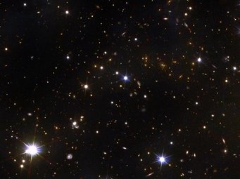 Ein Schnappschuss mit Tiefgang: Dieser Bildausschnitt, der etwa 200 Mal kleiner ist als die Gesamtaufnahme des Perseus-Clusters, gibt einen Eindruck der Details, die neben dem prächtigen Bild des Perseus Clusters im Vordergrund untergehen. Die hellsten Punkte mit sechs sternförmig angeordneten „Spikes“ sind Sterne unserer Galaxie im Vordergrund. Dazwischen liegen zahlreiche diffuse und rötliche Flecken, die Galaxien aus der Frühzeit des Universums entsprechen. Manche sind so weit entfernt, dass ihr Licht 10 Milliarden Jahre gebraucht hat, um uns zu erreichen. Weißliche Punkte mit sechs sternförmig angeordneten Spitzen vor schwarzem Hintergrund, dazwischen zahlreiche schwach erkennbare, kleine ovale rötliche Flecken