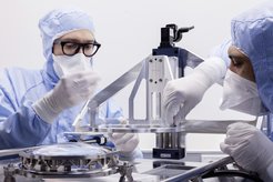 Nachaufnahme von zwei Personen in OP Bekleidung und FFP Masken, die an einer hell metallen glänzenden Apparatur hantieren. Links im Bild befindet sich eine optische Linse, etwa handtellergroß, eingefasst in einem metallenen Rahmen, rechts im Bild das Metallgerippe, auf das das Linsensystem aufgesetzt werden soll.