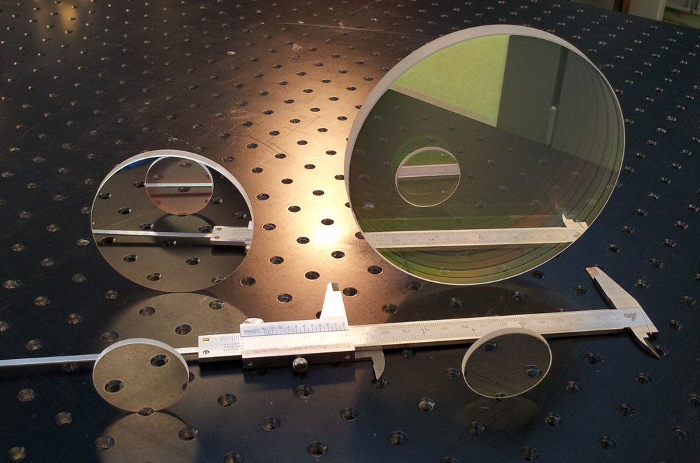 The four infrared filters installed in the NISP instrument with diameters of four centimetres in the foreground and eight and fourteen centimetres in the background. At over 300 grams per filter, these are the largest and heaviest infrared filters ever used on an astronomical space mission. The basis of all filters is quartz glass, which is specially coated so that each filter only allows infrared light at very specific wavelengths to pass through. Two small circular discs of the same size stand upright next to each other on a perforated plate, directly behind them in a second row are a disc about twice as large and a disc four times as large. A calliper lies between the two rows. The discs reflect what is in front of them.