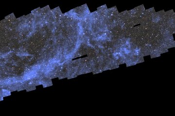 A wide, elongated black background with a long blueish patched-up field of stars, galaxies, and wispy clouds in the upper left diagonal of the image. This patchy mosaic consists of multiple almost square photographs arranged in a shape that loosely resembles a thin, pixelated whale diving towards the bottom left of the image. The myriad of sparkling galaxies and stars in this mosaic crowd together to create the impression of a dark grey field with frequent speckles of bright pastel yellow, red and blue dots of light at an even distribution. A streak of silver-blue diffuse clouds in a half-elliptical shape in the left half of the image and a small round patch just right of the centre stand out particularly. There are small rectangular holes in the mosaic at three spots right of the image centre. The pitch-black background is peaking through and starkly contrasts the sparkling image field.