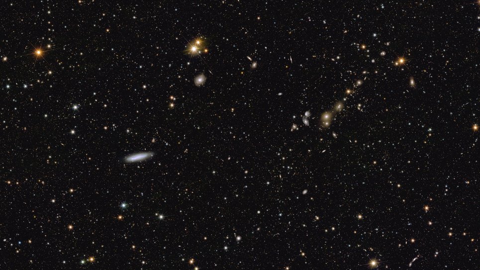 Ein Meer von Lichtpunkten, die gleichmäßig über einen schwarzen Hintergrund verstreut sind, wobei einige hellere und bunte Punkte hervorstechen. Wenn man von links nach rechts über das Bild blickt, fällt in der linken oberen Ecke zuerst ein heller orangefarbener Stern mit sechs schwachen Zacken ins Auge. Unten rechts davon, knapp unterhalb der Bildmitte, schwebt eine dünne weiße elliptische Galaxie ins Bild. Etwas weiter rechts darüber tanzt ein Paar heller goldgelber Sterne ins Bild. Weiter rechts, im letzten Drittel des Bildes, ist eine kurze Reihe von Sternen und Galaxien zu sehen. Diese erscheint wie eine im 45-Grad-Winkel geneigte Kette, an der dunstige Galaxien in verschiedenen Gelbtönen wie Perlen an einem kosmischen Armband aufgereiht sind.