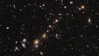 This image shows an area of the mosaic released by ESA’s Euclid space telescope on 15 October 2024. The area is zoomed in 36 times compared to the large mosaic. In this image, the core of galaxy cluster Abell 3381 is visible, 678 million light-years away from us. The image shows many different galaxies of various shapes and sizes, from massive elliptical to modest spiral galaxies, down to tiny and dim dwarf galaxies. A dark black backdrop with bright dots of stars and disks of galaxies scattered across it. Fading into the background are myriad light points, like specks of white paint and light hues of yellow, blue, and purple. In the foreground, a handful of bright light sources draw attention. Spanning from the top right corner to the bottom left of the image, yellow light points form a diagonal string of sparkling beads. Some of these are sharp, piercing dots with six faint spikes; others are blurry blobs of light, gleaming bright in their centre with their edges blending into the depths of the black background. In the lower left quarter of the image, two hazy white spiral shapes of light catch the eye, which appear to be swirling around each other.