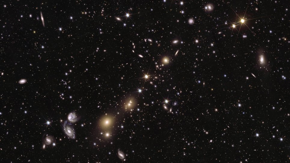 Ein dunkler schwarzer Hintergrund mit hellen Sternenpunkten und Galaxienscheiben, die über den Hintergrund verstreut sind. Im Hintergrund sind unzählige Lichtpunkte zu sehen, wie weiße Farbflecken und helle Farbtöne in Gelb, Blau und Violett. Im Vordergrund ziehen eine Handvoll heller Lichtquellen die Aufmerksamkeit auf sich. Gelbe Lichtpunkte, die sich von der rechten oberen Ecke bis zur linken unteren Ecke des Bildes erstrecken, bilden eine diagonale Kette aus funkelnden Perlen. Einige von ihnen sind scharfe, stechende Punkte mit sechs schwachen Zacken; andere sind verschwommene Lichtkleckse, die in ihrer Mitte hell leuchten und deren Ränder in die Tiefe des schwarzen Hintergrunds übergehen. Im unteren linken Viertel des Bildes fallen zwei verschwommene weiße Lichtspiralen ins Auge, die sich umeinander zu drehen scheinen.