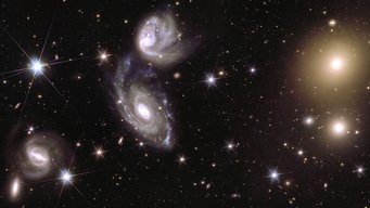 This image shows an area of the mosaic released by ESA’s Euclid space telescope on 15 October 2024. The area is zoomed in 150 times compared to the large mosaic. On the left of the image, Euclid captured two galaxies (called ESO 364-G035 and G036) interacting with each other, 420 million light-years from us. On the right of the image, galaxy cluster Abell 3381 is visible, 678 million light-years away from us. Three groups of light sources and a scatter of piercing dots of light with six faint spikes stand out in stark contrast against a black backdrop. The most prominent light sources occupy the centre of the image. They are two hazy white spirals, that appear to be swirling in a cosmic dance with each other, with the lower spiral being larger than the one above it. On the right side of the image, two spots of gleaming yellow light draw attention. The hazy light blobs emit a golden glow from their centre, which fades out in a circular shape into the background. Another spiral shape can be seen in the bottom left corner of the picture. It appears as if it is a thin white bar spinning in a circle and emitting a white spray of paint at its ends, leaving behind a diffuse trace of light.