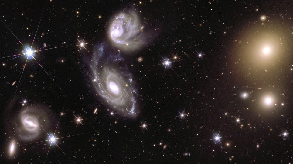 Drei Gruppen von Lichtquellen und eine Streuung von durchdringenden Lichtpunkten mit sechs schwachen Zacken heben sich in starkem Kontrast von einem schwarzen Hintergrund ab. Die auffälligsten Lichtquellen befinden sich in der Mitte des Bildes. Es handelt sich um zwei dunstige weiße Spiralen, die in einem kosmischen Tanz miteinander zu tanzen scheinen, wobei die untere Spirale größer ist als die darüber liegende. Auf der rechten Seite des Bildes ziehen zwei Flecken mit schimmerndem gelbem Licht die Aufmerksamkeit auf sich. Die dunstigen Lichtkleckse strahlen aus ihrem Zentrum ein goldenes Leuchten aus, das kreisförmig im Hintergrund verschwindet. Eine weitere Spiralform ist in der linken unteren Ecke des Bildes zu sehen. Es scheint ein dünner weißer Balken zu sein, der sich im Kreis dreht und an seinen Enden einen weißen Farbnebel abgibt, der eine diffuse Lichtspur hinterlässt.