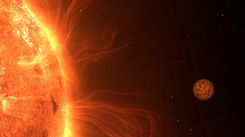 An artistic impression of Trappist-1 b shortly before it passes behind the cool, red dwarf star, Trappist-1. Such stars are known for their activity with large starspots and eruptions. Trappist-1 b may experience intense volcanism. A close-up of a star dominates the left side of the image. Its surface glows intensely in various shades of orange, showing irregular structures like darker spots. Delicate, glowing plasma filaments arc outward from the Sun's surface. On the right side of the image, a small rocky planet is visible, its reddish-brown surface marked with dark, mottled patterns. The background is black, dotted with a few white star-like points.