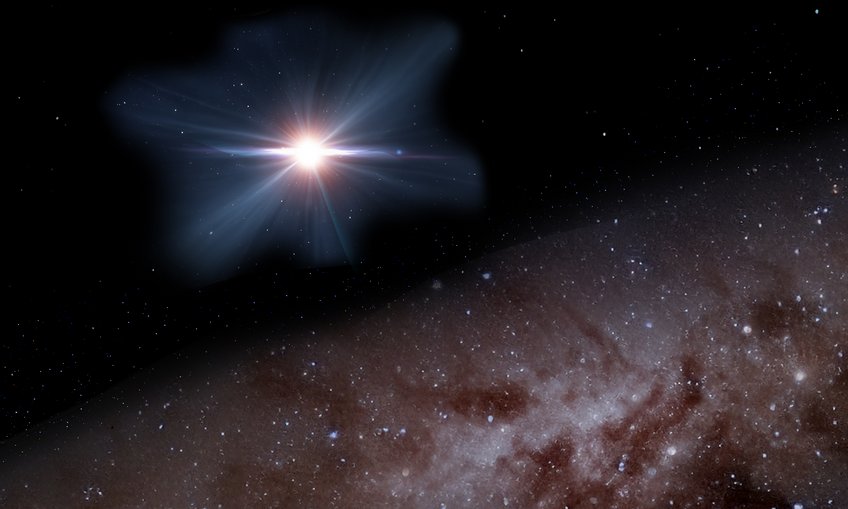 Artist’s impression of the bright, very early active galactic nucleus that was found by Bañados and his colleagues, which has fundamental implications for black hole growth in the earliest billion or so years of cosmic history. The image depicts an artistic rendering of a bright blazar, a supermassive black hole at the center of a distant galaxy, ejecting high-energy matter in the form of jets into space. The blazar appears as a glowing point of light with bluish-white rays scattering in various directions. In the background, a field of stars is visible, while the lower part of the image shows a portion of the Milky Way or another galaxy with brown and white gradients.