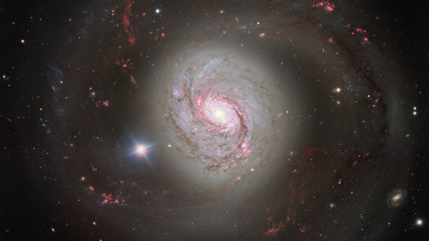 An image of the spiral galaxy NGC 1068 (Messier 77) obtained by the European Southern Observatory’s (ESO) Very Large Telescope (VLT). The galaxy has a distance of 14.4 Mpc (47 million light-years) and is one of the nearest galaxies with an active galactic nucleus. A vivid image of the spiral galaxy Messier 77 (M77), also known as NGC 1068. The galaxy exhibits an impressive structure with a brightly glowing centre and spiralling arms extending far into space. The spiral arms are highlighted by pink and red regions, representing areas of active star formation. The galaxy is surrounded by countless small points of light, representing distant stars and galaxies. In the foreground, a bright, star-like point shines on the left side of the galaxy. The galaxy's colours range from white and blue in the centre to reddish hues in the outer regions.