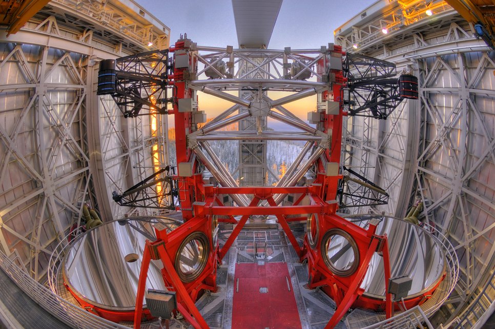 Das Bild zeigt das Innere des Large Binocular Telescope (LBT), einem der weltweit leistungsstärksten Teleskope. Im Zentrum befinden sich zwei große, runde, stark reflektierende Hauptspiegel, die parallel nebeneinander angeordnet sind. Die Spiegel sind von einer massiven, geometrisch komplexen roten Stahlkonstruktion umgeben, die durch diagonale und rechtwinklige Verstrebungen Stabilität gewährleistet. Über den Spiegeln ragen zwei schwarze Armstrukturen mit zylindrischen Komponenten heraus, die vermutlich Instrumente oder Sensoren enthalten. Das Dach des Teleskops ist geöffnet, wodurch die umliegende, symmetrische Metallstruktur mit ihren bogenförmigen und rechteckigen Elementen sichtbar wird. Im Hintergrund erstreckt sich eine schneebedeckte Waldlandschaft unter einem Himmel, der in sanftem Orange- und Blauton leuchtet. Die Szene kombiniert technische Präzision mit ästhetischer Harmonie und bietet einen beeindruckenden Einblick in die geometrische Eleganz dieses Forschungsinstruments.