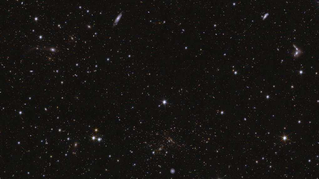 This image shows an area of Euclid’s Deep Field South. The area is zoomed in 16 times compared to the large mosaic. Many galaxies are visible in this field, all with different shapes and colours because they have different ages and distances. A sea of stars and galaxies sparkle against a black background. Several galaxies can be identified by their elongated shape and/or spiral arms. Some galaxies are seen edge-on while one prominent spiral galaxy at the bottom centre is seen face-on. At the far right, between the middle and top of the image, are some interacting galaxies. Galaxy clusters are also seen, in particular near bottom centre, where features smeared into arcs represent gravitational lensing. The brightest stars in the image have diffraction spikes.