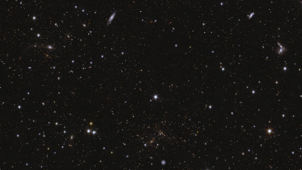A sea of stars and galaxies sparkle against a black background. Several galaxies can be identified by their elongated shape and/or spiral arms. Some galaxies are seen edge-on while one prominent spiral galaxy at the bottom centre is seen face-on. At the far right, between the middle and top of the image, are some interacting galaxies. Galaxy clusters are also seen, in particular near bottom centre, where features smeared into arcs represent gravitational lensing. The brightest stars in the image have diffraction spikes.