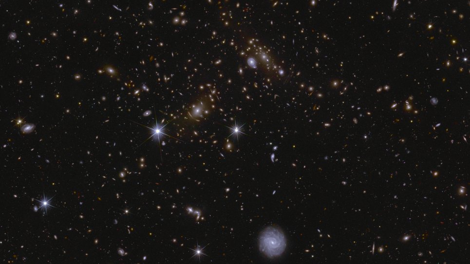 A sea of galaxies of many different shapes and sizes, oriented in all directions and many with spiral arms visible. A large, nearby face-on spiral galaxy draws attention at the bottom centre of the image. Two bright stars in the centre and to the left are seen with prominent diffraction spikes. In between the two stars lie a lensed galaxy cluster, with arc-like smears surrounding the central bright galaxy cluster.
