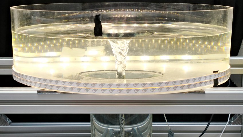 This photo shows the upper part of the water tornado model. The acrylic glas tank has a diameter of 50 centimetres and is illuminated with LED strips. The water forms a vortex whose surface shape reproduces the profile of a gravitational field. The analysis showed that the motion of the water closely resembles the behaviour seen in protoplanetary discs. A cylindrical, transparent container stands on an open, silver-coloured metal frame. It is filled with clear water, at the centre of which a slender, funnel-shaped vortex rotates from surface to bottom. The water surface is indented at the centre and slightly rippled at the edges. Tiny air bubbles are drawn downward by the vortex, forming fine lines along the inner wall. LED light strips encircle the container, making the vortex stand out starkly against the dark surroundings. The scene has the appearance of a scientific demonstration visualising fluid dynamics.