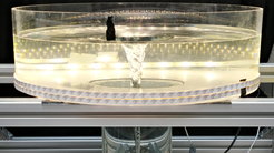 A cylindrical, transparent container stands on an open, silver-coloured metal frame. It is filled with clear water, at the centre of which a slender, funnel-shaped vortex rotates from surface to bottom. The water surface is indented at the centre and slightly rippled at the edges. Tiny air bubbles are drawn downward by the vortex, forming fine lines along the inner wall. LED light strips encircle the container, making the vortex stand out starkly against the dark surroundings. The scene has the appearance of a scientific demonstration visualising fluid dynamics.