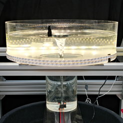 Photograph of the water tornado model in a darkened laboratory. At the centre stands a silver-coloured metal structure supporting a circular, transparent acrylic glass tank with a diameter of 50 cm. The outer wall of the tank is lined with narrow white LED strips, emitting an even glow from bottom to top. Beneath the wide tank is a narrower cylinder, whose smooth walls extend down to two black nozzles. Hoses are attached to these nozzles, continuously feeding water into the system. The inflow of water produces a slender, funnel-shaped vortex stretching from the surface to the bottom and visually resembling a gravitational field in profile. Several cables and hoses run along the metal frame. The bright water vortex stands out starkly against the dark background, conveying the impression of a precisely controlled scientific experiment.