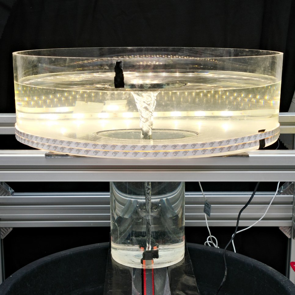 Fotografie eines Wasser-Tornado-Modells in einem abgedunkelten Labor. Im Zentrum steht eine silberfarbene Metallkonstruktion, auf der ein kreisrundes, transparentes Plexiglas-Becken mit 50 cm Durchmesser ruht. Die Außenwand des Beckens ist mit schmalen weißen LED-Streifen versehen, die von unten nach oben gleichmäßig leuchten. Unter dem breiten Becken schließt sich ein engerer Zylinder an, dessen glatte Wände nach unten bis zu zwei schwarzen Düsen führen. An diesen Düsen sind Schläuche angeschlossen, die kontinuierlich Wasser einspeisen. Durch den Wasserzufluss bildet sich ein schlanker, trichterförmiger Wirbel, der von der Oberfläche bis zum Boden reicht und im Profil an ein Gravitationsfeld erinnert. Mehrere Kabel und Schläuche verlaufen entlang der Metallkonstruktion. Der helle Wasserwirbel hebt sich kontrastreich vom dunklen Hintergrund ab und vermittelt den Eindruck eines präzise kontrollierten wissenschaftlichen Experiments.