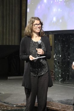 Eine Frau, nämlich Ruth Grützbauch, steht lächelnd auf einer Bühne. Sie hält eine Medaille und ein Stück Papier in der Hand.