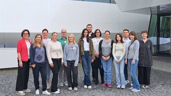 Teilnehmende am Willkommenstag der Auszubildenden. Von links nach rechts: Simone Rosteck (MPIA), Anna Michalczyk-Schneider (MPIL), Kristine Pöhner (MPIK), Leonie Barth (Auszubildende MPIL), Katrin Hohmeyer (MPIK), Lilli Schmitt (Auszubildende MPIK), Lara Dinkic (Auszubildende MPIMR), Jakob Schatz (Auszubildender MPIA), Annika Kluth (Auszubildende MPIA), Lisa Mirus (MPIA), Larissa Nachmann (Auszubildende MPIMR), Jörg Graf (MPIMR), Julia Gauglitz (MPIMR), Emma Sofie Krojanski (FJN MPIA). Gruppenfoto mit 14 freundlich blickenden Personen, die sich in zwei Reihen aufgestellt haben. Sie stehen auf einer Fläche mit grauen Pflastersteinen. Im Hintergrund ist die weiße Fassade des Hauses der Astronomie zu sehen, etwa 5 Meter entfernt.
