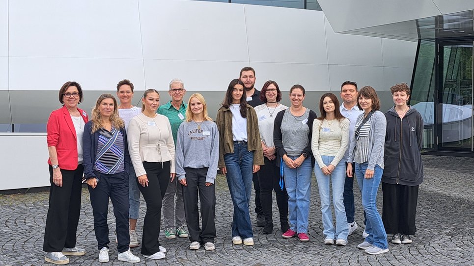 Group photo with 14 people smiling, arranged in two rows. They are standing on a surface of grey paving stones. In the background, about five metres away, the white façade of the Haus der Astronomie is visible.