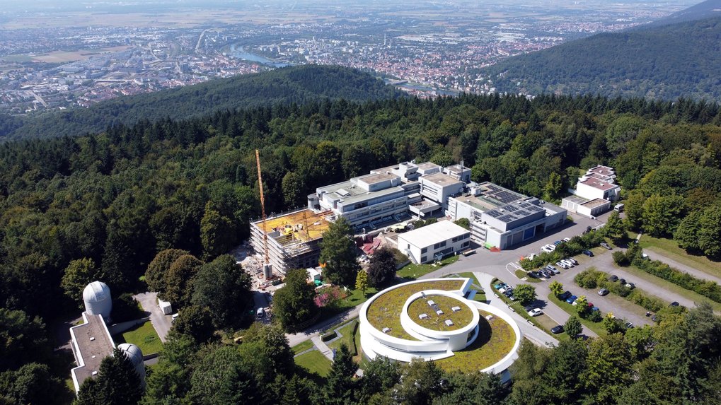 Aerial view of the MPIA Campus. Aerial view of the campus of the Max Planck Institute for Astronomy (MPIA) on the Königstuhl hill in Heidelberg. In the foreground at the lower right stands the Haus der Astronomie, a modern white building in a spiral shape with a green roof. To its left are several additional institute buildings with flat roofs, some fitted with solar panels, and a new construction site with a crane and yellow scaffolding. In the lower left, two white domes of small observatories are visible. Parking areas with cars are located in front of the buildings. The entire campus is surrounded by dense green forest. In the background stretches the Neckar valley with the city of Heidelberg, the river with bridges, and the wide Upper Rhine Plain extending to the horizon.