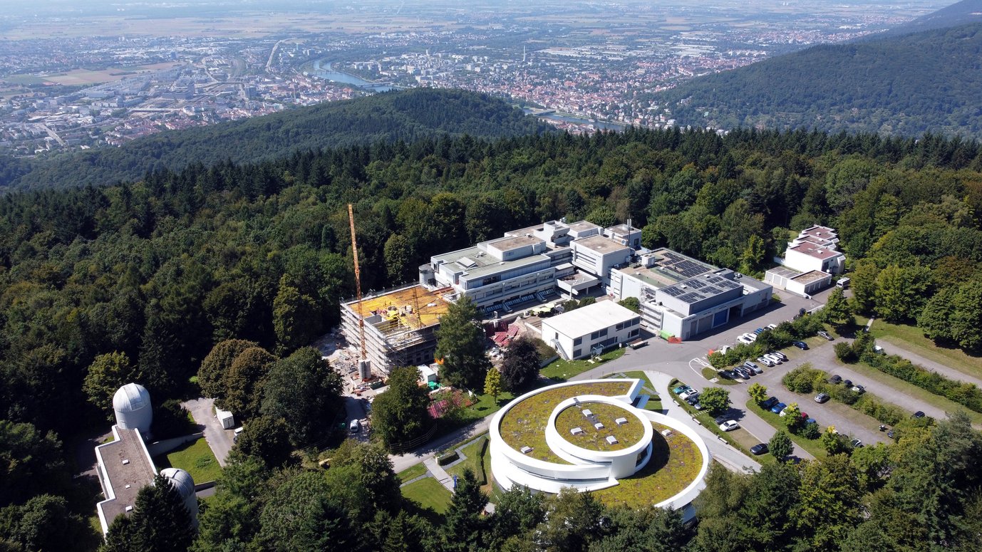 Aerial view of the campus of the Max Planck Institute for Astronomy (MPIA) on the Königstuhl hill in Heidelberg. In the foreground at the lower right stands the Haus der Astronomie, a modern white building in a spiral shape with a green roof. To its left are several additional institute buildings with flat roofs, some fitted with solar panels, and a new construction site with a crane and yellow scaffolding. In the lower left, two white domes of small observatories are visible. Parking areas with cars are located in front of the buildings. The entire campus is surrounded by dense green forest. In the background stretches the Neckar valley with the city of Heidelberg, the river with bridges, and the wide Upper Rhine Plain extending to the horizon.