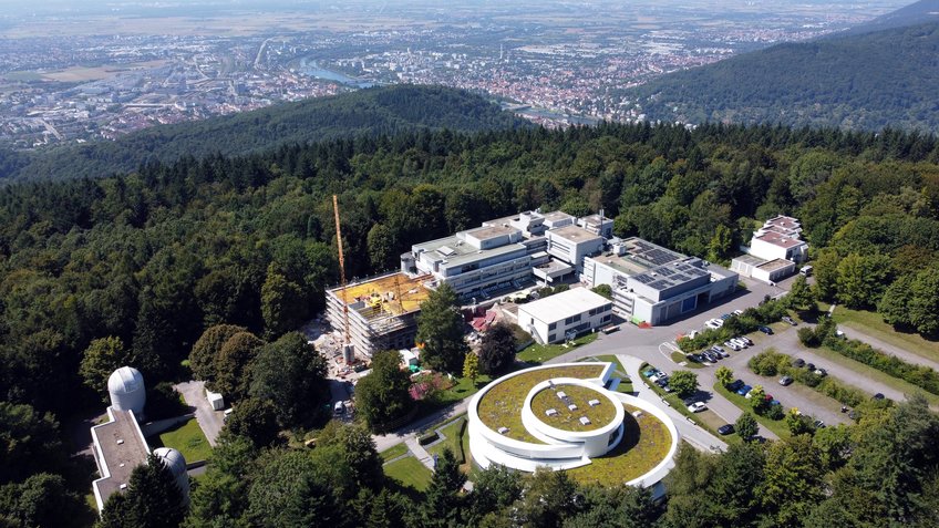 Luftbild des MPIA-Campus. Luftaufnahme des Campus des Max-Planck-Instituts für Astronomie (MPIA) auf dem Königstuhl in Heidelberg. Im Vordergrund rechts unten ist das Haus der Astronomie zu sehen, ein modernes, weißes Gebäude in Spiralform mit begrüntem Dach. Links davon befinden sich mehrere weitere Institutsgebäude mit Flachdächern, teilweise mit Solarpaneelen, sowie ein Neubau mit Baukran und gelber Gerüstverkleidung. Im unteren linken Bereich sind zwei weiße Kuppeln kleiner Observatorien zu erkennen. Vor den Gebäuden liegen Parkplätze mit geparkten Autos. Umgeben ist der Campus von dichtem grünen Wald. Im Hintergrund erstreckt sich das Neckartal mit der Stadt Heidelberg, Flussläufen, Brücken und dem weiten, flachen Oberrheingebiet bis zum Horizont.