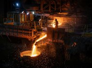 In einer dunklen Halle fließt hell glühendes Metall aus einem Ofen in eine runde mannshohe Wanne. Vor dem Ofen steht ein Mann mit Schutzhelm.
