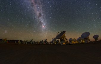 The Atacama Large Millimeter/submillimeter Array (ALMA) on the Chajnantor Plateau in Chile at night. Night-time view of the Atacama Large Millimeter/submillimeter Array (ALMA) in the Atacama Desert: many large parabolic antennas stand on a wide, flat high-altitude plateau, arranged in rows. Above the observatory stretches a clear, star-filled sky crossed by the bright band of the Milky Way. The antennas are faintly illuminated and appear as dark silhouettes and pale dishes against the sky, emphasising the scale of the facility and the remote, light-pollution-free environment.