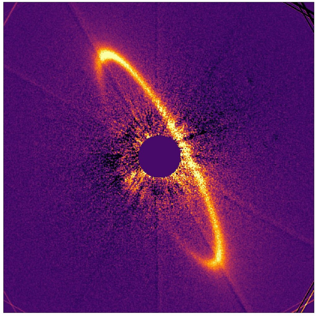 Falschfarbenaufnahme einer Trümmerscheibe um einen Stern, aufgenommen mit dem Instrument SPHERE. In der Bildmitte ist der Stern durch eine runde, dunkle Maske abgedeckt. Um ihn herum ist eine helle, gebogene Staubstruktur zu sehen, die einen asymmetrischen, bogenförmigen Teil einer Scheibe nachzeichnet. Die Scheibe erscheint in leuchtenden Gelb- und Orangetönen vor violettem Hintergrund, was die Verteilung von Staub und Streulicht sichtbar macht. Helligkeitsunterschiede und Unregelmäßigkeiten deuten auf eine verzerrte oder dynamisch beeinflusste Struktur der Trümmerscheibe hin.