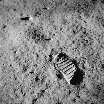 Eine Nahaufnahme des Stiefelabdrucks eines Astronauten im feinpulvrigen lunaren Regolith während der Außenbordaktivitäten (EVA) von Apollo 11 auf dem Mond. Ähnliche Bedingungen könnten auf dem Exoplaneten LHS 3844 b herrschen, bedingt durch langanhaltende Weltraumverwitterung infolge der stellaren Einstrahlung und Meteoriteneinschläge. Eine Schwarz-Weiß-Nahaufnahme des Stiefelabdrucks eines Astronauten im feinen, grauen Regolith einer fremden Oberfläche. Der Abdruck zeigt ein markantes, geripptes Profil, das tief in den staubigen Boden eingedrückt ist und von kleinen Kieselsteinen übersät ist. Harte Schatten betonen die Textur des Untergrunds. Der Boden besteht aus einer dicken Schicht feinkörnigen, dunklen Staubs und kleinen Gesteinstrümmern. Der im unteren rechten Quadranten positionierte Abdruck ist scharf und klar definiert; flach einfallendes Licht erzeugt tiefe Schatten innerhalb der Profilrillen und hinter kleinen Oberflächenfelsen. Dies unterstreicht die Konsistenz des Bodens in einer Welt ohne Wind oder Atmosphäre, in der solche Strukturen erodieren könnten.