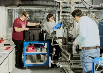 Am Max-Planck-Institut für Astronomie schützt sich Klaus Paschek (rotes T-Shirt) vor einem UV-Laser, der künstlich erzeugte Moleküle anregt, wie sie im All entstehen könnten. Michael Hermann (Mitte) und Tushar Suhasaria (vorne) treffen Vorbereitungen, um diese Moleküle zu analysieren. Labor mit drei Personen und technischer Ausrüstung.