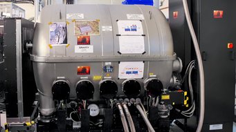 The GRAVITY instrument of the Very Large Telescope Interferometer (VLTI) at the European Southern Observatory’s (ESO) Paranal Observatory. The focus is on the Gravity spectrograph, an instrument at ESO's Paranal Observatory and part of the VLTI. The device features a large, silver cylindrical main chamber with multiple attached cables and pipes. The front displays four round ports with labels and measuring instruments. Several printed sheets and photos, including one labelled 'GRAVITY', are attached to the chamber's surface. Numerous pipes and cables run along the wall in the background. On the right, a tall, dark control cabinet is connected by additional cables.
