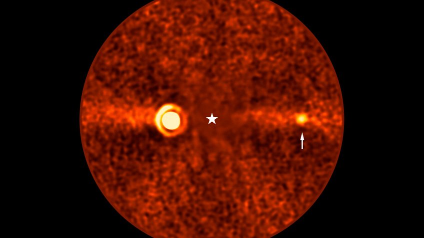 Diese Aufnahme zeigt den neu entdeckten Planeten Beta Pictoris d, der den Stern Beta Pictoris umkreist. Die Position des Sterns ist in der Mitte des Bildes markiert; sein Licht wurde jedoch während der Datenverarbeitung herausgerechnet, um die unmittelbare Umgebung sichtbar zu machen. Der neue Planet ist mit einem Pfeil gekennzeichnet und der dritte, der um diesen Stern nachgewiesen werden konnte. Bei den beiden anderen handelt es sich um Beta Pictoris b – das helle Signal auf der linken Seite – und Beta Pictoris c, der seine Umlaufbahn wesentlich näher am Stern hat und auf diesem Bild nicht zu sehen ist. Das diffuse, waagerechte Band im Hintergrund zeigt die Trümmer- und Staubscheibe um den Stern in Kantenstellung (Edge-on-Ansicht), die aus den Überresten der Planetenentstehung besteht. Runde, rötlich gefärbte astronomische Aufnahme mit körniger Struktur. Im Zentrum befindet sich ein weißes Sternsymbol, das die Position des Sterns Beta Pictoris markiert, der auf dem Bild nicht sichtbar ist. Links vom Zentrum ist ein auffällig leuchtender Bereich mit konzentrischen Kreisen zu sehen, ein Abbild des Exoplaneten Beta Pictoris b. Eine horizontale, helle Linie durchzieht das Bild und repräsentiert die Trümmerscheibe um den Stern. Rechts vom Zentrum hebt ein kleiner, heller Punkt, markiert durch einen weißen Pfeil, einen weiteren Exoplaneten hervor: Beta Pictoris d.