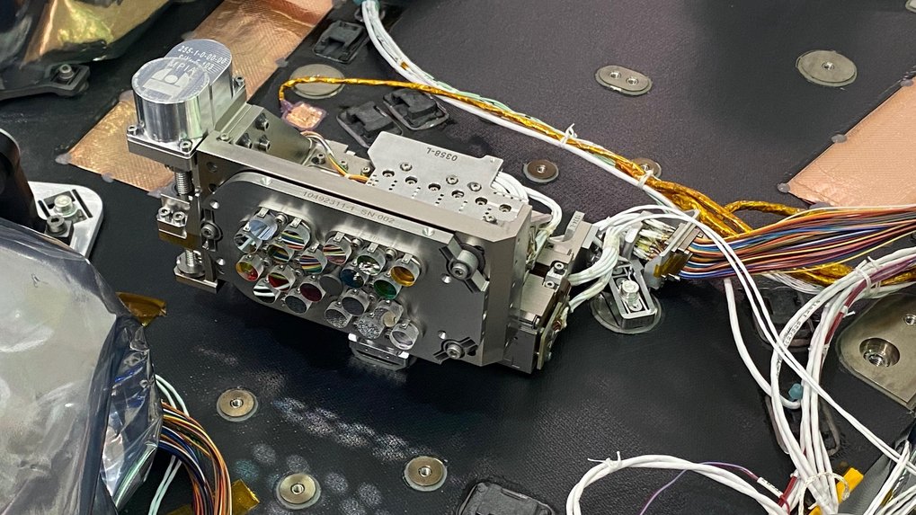A precision alignment mechanism is mounted on an optical bench. Several optical filters are integrated into the metallic housing, which features multiple openings and screw connections. Numerous coloured and white cables are attached to the device and run neatly across the surface of the optical bench. The setting appears technical and organized.