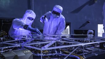 Fully populated optical bench of the CGI following delivery to NASA's Goddard Space Flight Center. At its maximum dimension, the optical bench measures 1.7 metres × 1.7 metres. An optical bench with various electronic components and cables is positioned in the foreground. Two individuals wearing full cleanroom attire, including coveralls, gloves, and protective eyewear, are working intently on the optical bench. The scene is illuminated with blue light, highlighting the technical environment and cleanroom setting. The individuals are touching or adjusting parts on the optical bench.