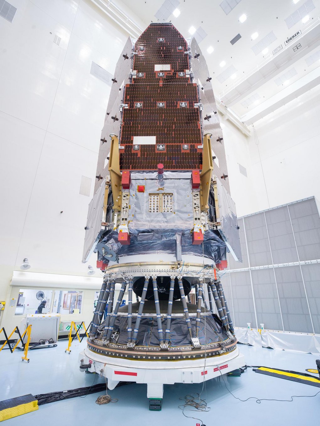 Ein großes Weltraumteleskop-Modul steht senkrecht in einer hell ausgeleuchteten Montagehalle. Das Modul ist an einem Adapter befestigt, der zahlreiche Verstrebungen und technische Komponenten aufweist. Die Nutzlast besteht aus einem länglichen, mit dunklen Paneelen bedeckten Hauptkörper, der nach oben in eine spitz zulaufende Verkleidung übergeht. Im Hintergrund sind Wände, Deckenpaneele und technische Einrichtungen der Halle zu sehen.