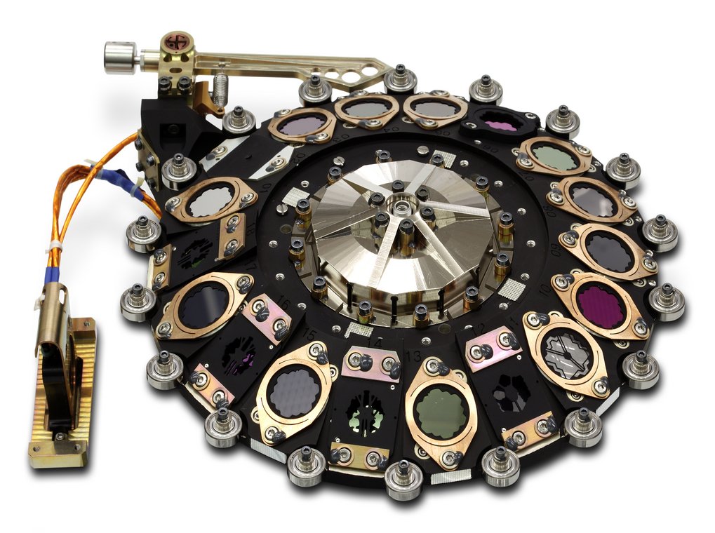 Ein hochpräzises mechanisches Filterrad für das MIRI-Instrument des James-Webb-Weltraumteleskops (JWST). Die kreisförmige schwarze Metalleinheit umfasst 18 ringförmig angeordnete Öffnungen. Einige Öffnungen enthalten lichtdurchlässige, farbige optische Filter, andere spezialisierte dunkle Masken für die Koronografie. Jeder Filter ist mit einer kupferfarbenen Halterung fixiert. Am Außenrand befinden sich kleine Kugellager aus Stahl für die Rotation. Links führt ein Bündel aus orangefarbenen und blauen Kabeln zu einem vergoldeten Elektronikbauteil. Am oberen Rand ist ein mechanischer Hebel- und Federmechanismus zur Positionierung erkennbar.
