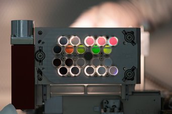Eine Ausführung des Precision Alignment Systems des Max-Planck-Instituts für Astronomie, das eine Serie Farbfilter in Position hält. Licht von der Gegenseite wird von jenen Filtern geblockt, die dunkel erscheinen. Der infrarote Teil des Lichts dringt durch die Filter, die rötlich erscheinen. Optisches Element mit kreisrunden Filtern unterschiedlicher Farbe